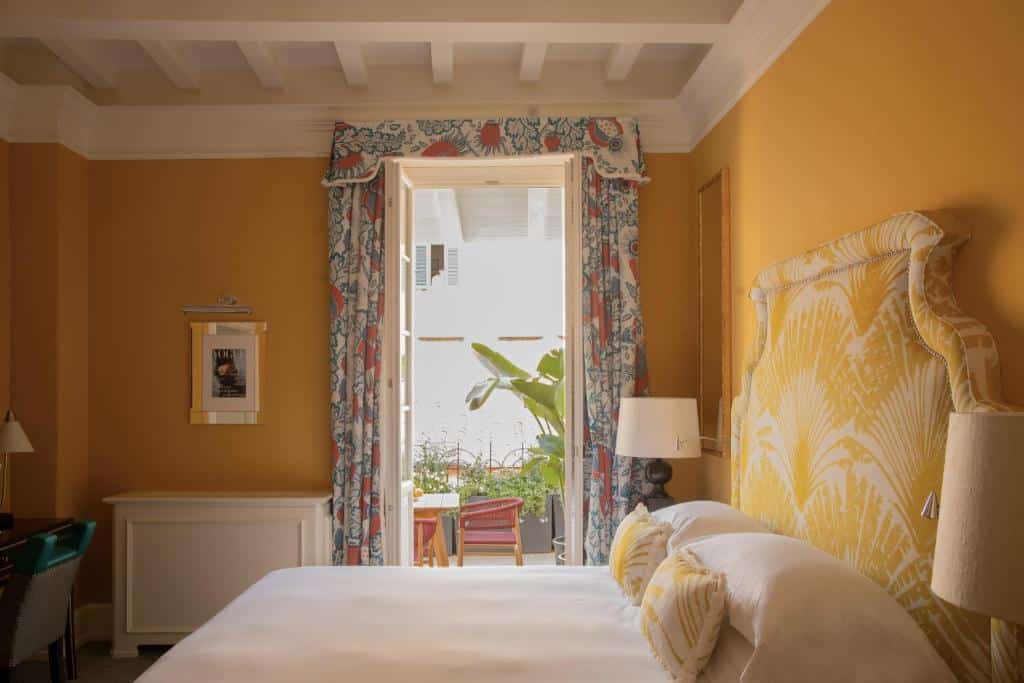 quarto do Maalot Roma, um dos hotéis perto da Fontana di Trevi, com cama de casal grande, mesa e candeeiro de cada lado, mesa e cadeiras na varanda, janela grande com cortinas floridas e paredes amarelas