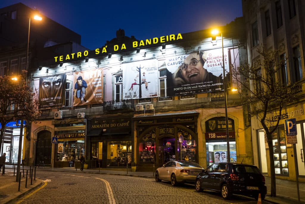 Fachada iluminada do Teatro Sá da Bandeira, no Porto, à noite, com cartazes e carros estacionados em frente. Representa o que fazer à noite no Porto.