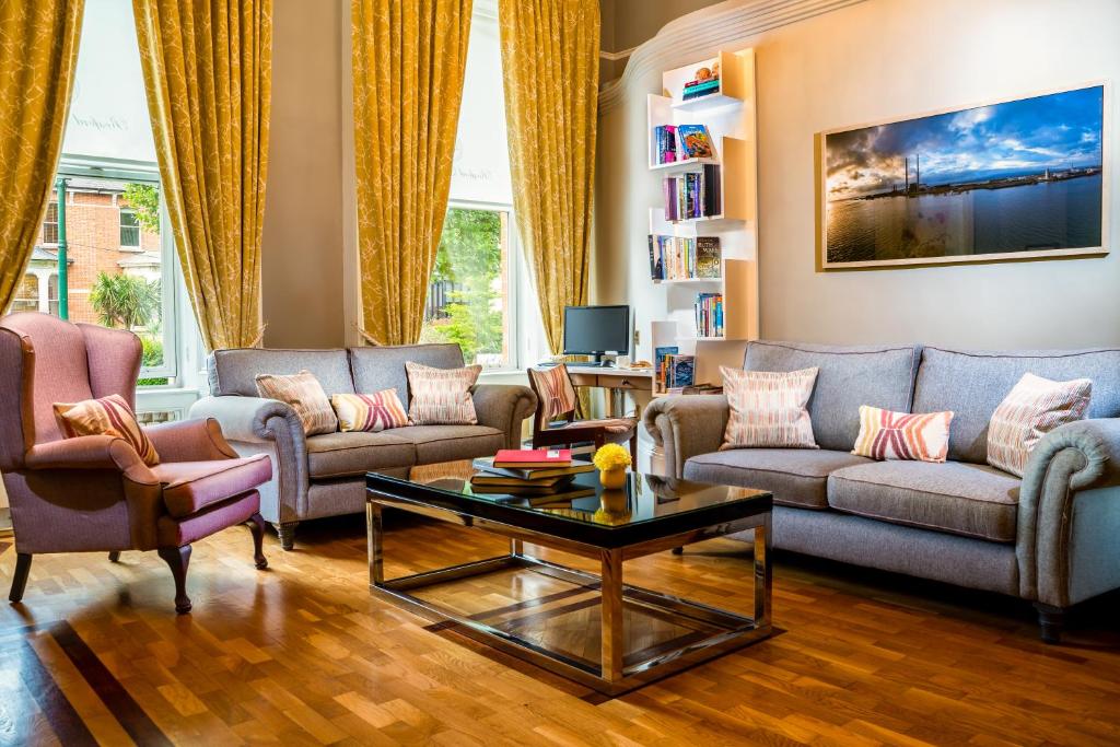 Sala de estar no Roxford Lodge Hotel com sofás cinzentos amplos, almofadas coloridas, estante com livros e televisão de ecrã grande com paisagem urbana ao fundo, e cortinas douradas.