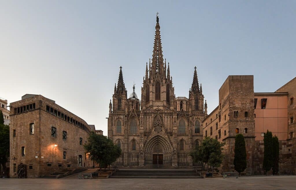 Entardecer junto à Catedral de Barcelona, uma igreja gótica com torres, rodeada de árvores e de outros edifícios antigos.
