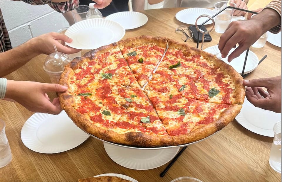 Várias mãos estendidas para pegar fatias de pizza sobre uma mesa de madeira, rodeada de pratos e copos.