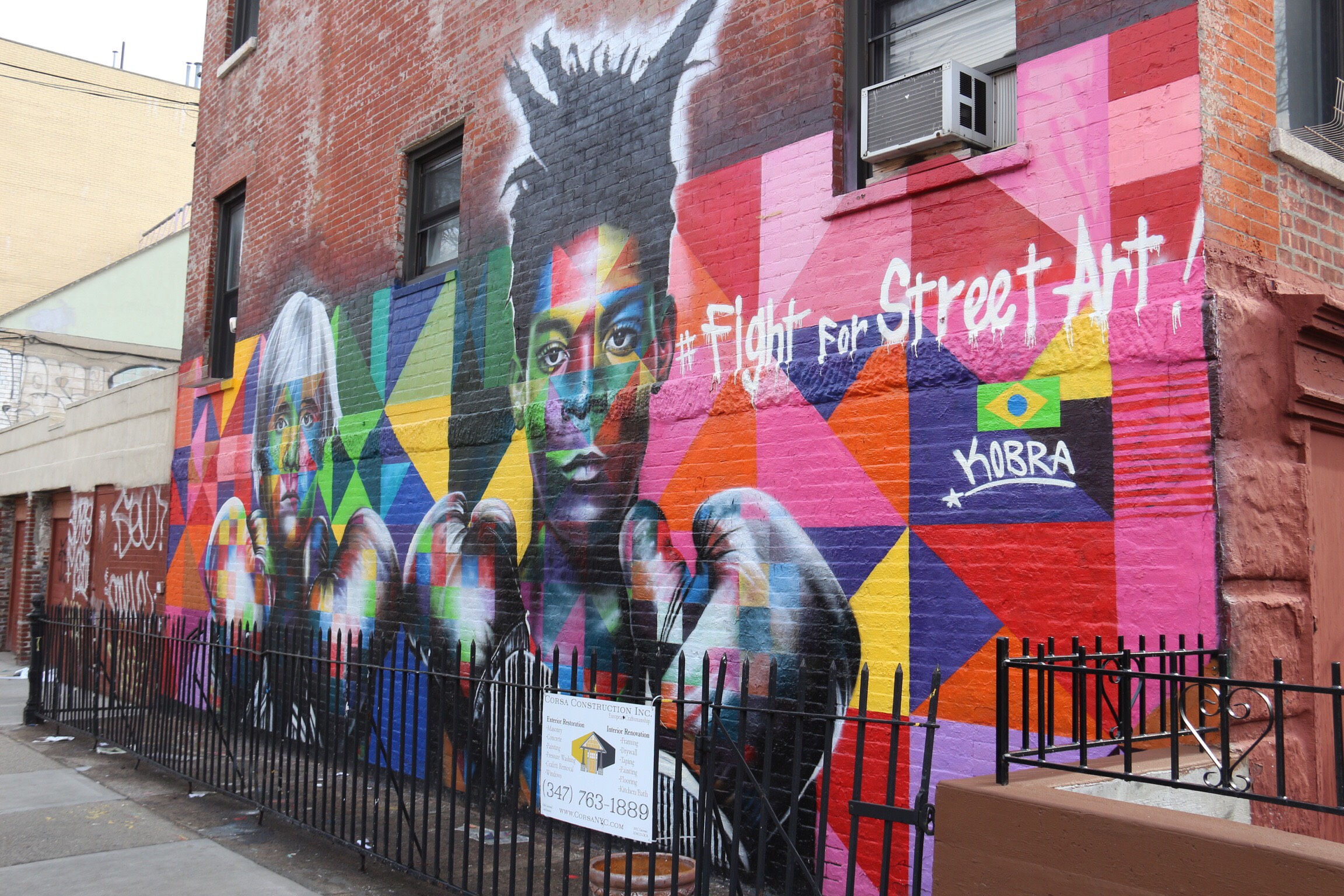 Grande mural colorido nas ruas de Williamsburg, a retratar um rosto com padrões geométricos vibrantes e um arco-íris ao fundo.