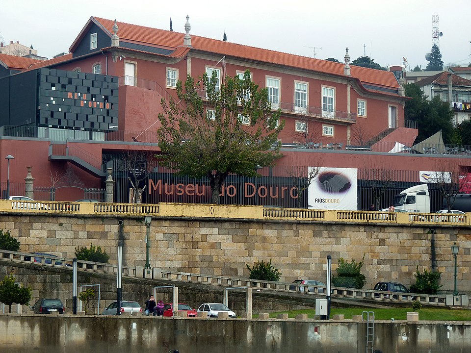 Edifício de tijolo vermelho que alberga o Museu do Douro, com carros estacionados ao longo de um muro de pedra.