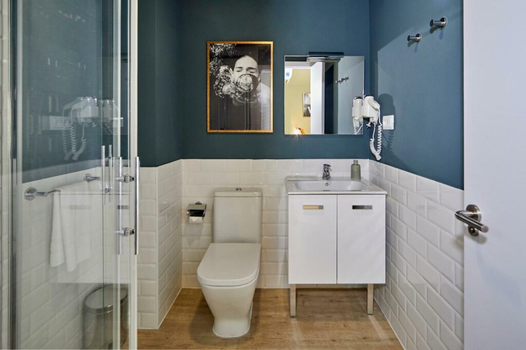 Casa de banho do L&H La Latina Selection. A sanita tem um lavatório com bancada ao lado e um quadro por cima. Na parede acima do lavatório há um espelho e um secador de cabelo. Em primeiro plano está um chuveiro com cabina e uma toalha branca pendurada num suporte ao lado.