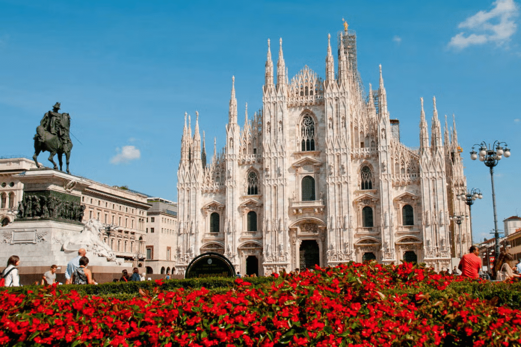 Vista do Duomo de Milão com muitas flores vermelhas em primeiro plano num jardim e uma escultura de um cavaleiro, com a imponente catedral neogótica de fachada branca ao fundo