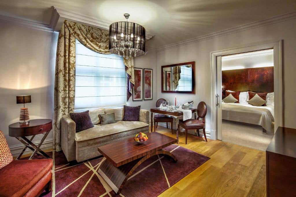Suíte elegante do The Grand Mark Prague, com sofá, mesa de centro, cadeiras e vista para um quarto com cama de casal.
