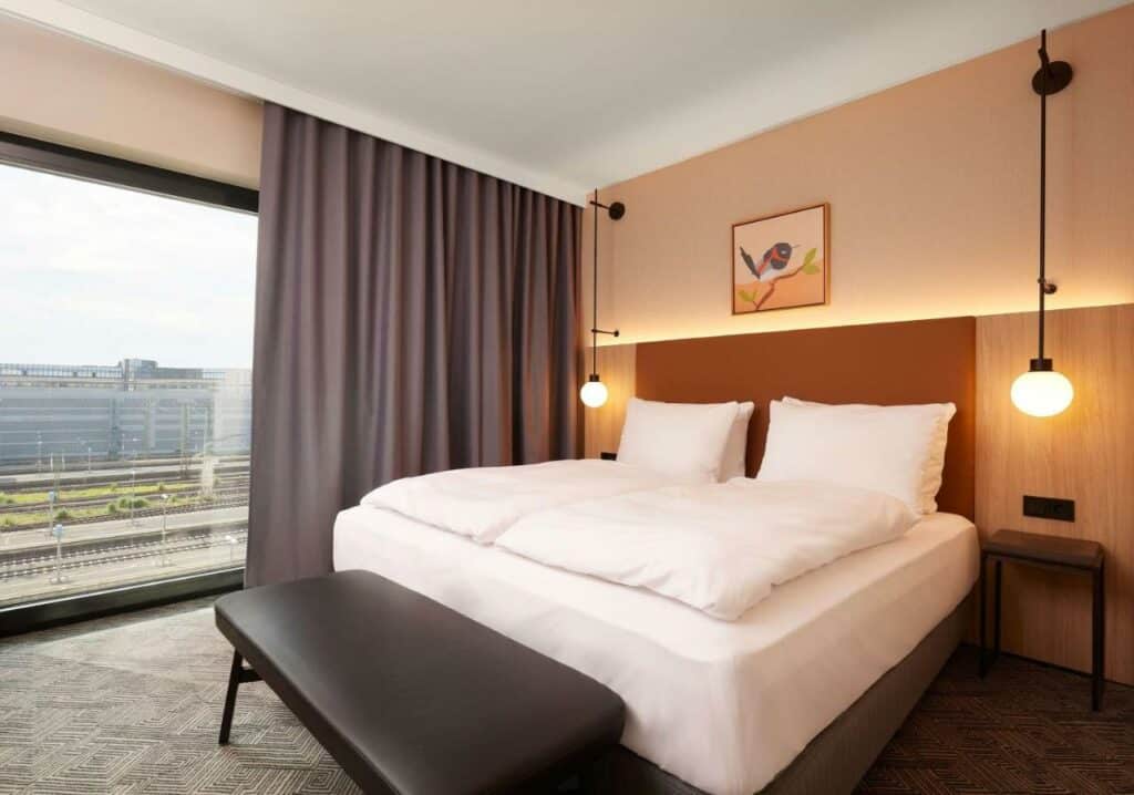 Quarto do Adina Apartment Hotel Dusseldorf com cama de casal e janelas panorâmicas com vista para a cidade. Representa hotéis em Dusseldorf.