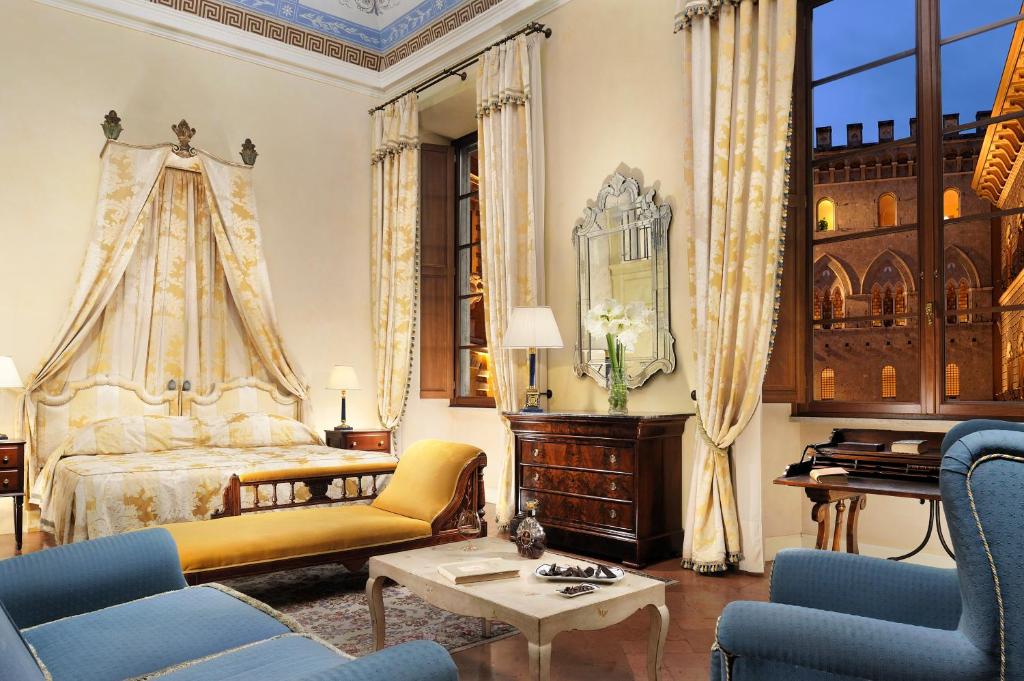 Suíte Heritage do Grand Hotel Continental Siena, de 50 m², com móveis de época, uma cama com colchas a combinar com as cortinas, poltronas grandes azuis, espelho e janelas com vista sobre um edifício semelhante a um castelo