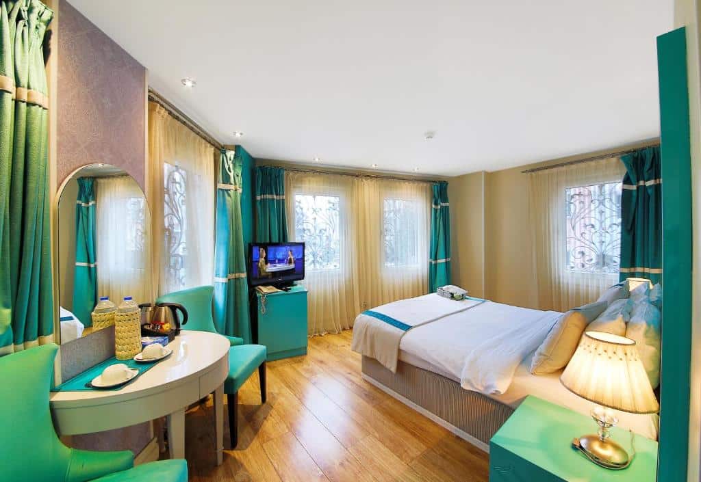 Um quarto do Katelya Hotel com uma cama de casal, detalhes em azul-petróleo, uma TV, uma secretária com duas cadeiras e janelas grandes com cortinados transparentes.