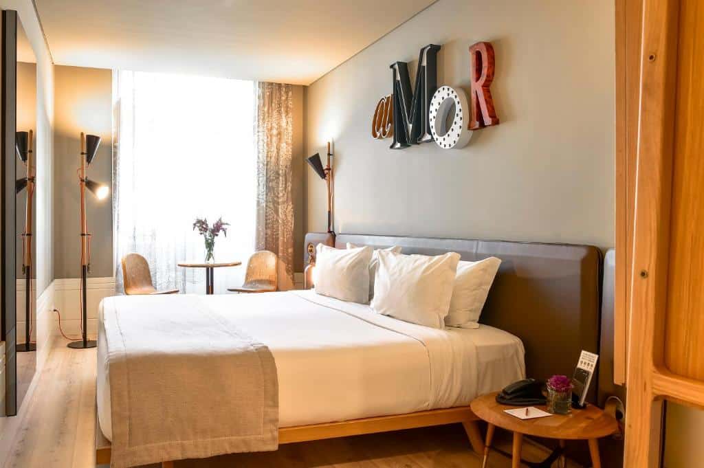 Quarto do My Story Hotel Rossio com cama de casal; ao fundo, uma janela com cortinas onde há uma pequena mesa e duas cadeiras; na parede em frente à cama, um espelho