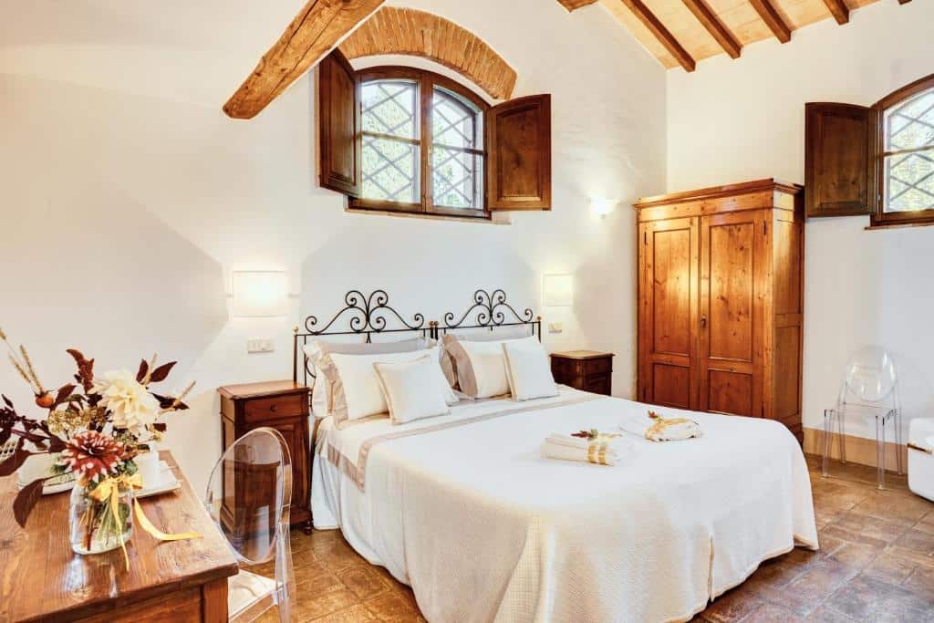 Quarto do Castello di Spedaletto da Laura, com 18 m², cama de casal com almofadas e toalhas enroladas em cima, mesa quadrada de madeira com um vaso de flores e cadeiras transparentes