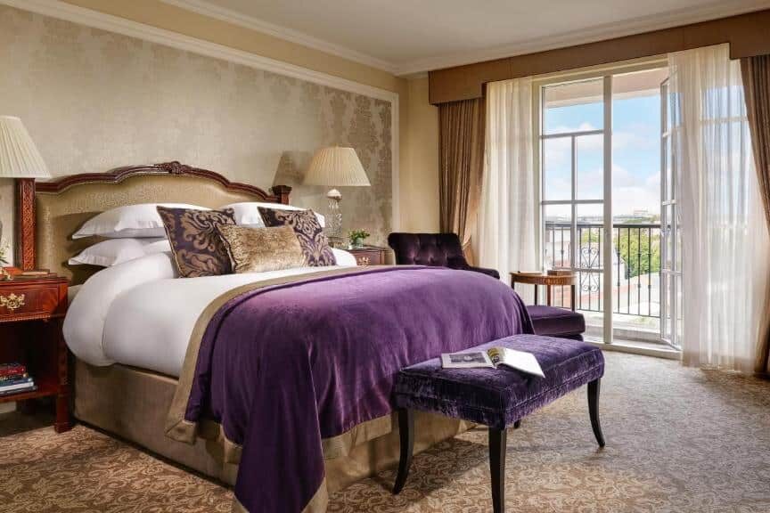 Quarto do InterContinental Dublin by IHG com cama de casal imponente, almofadas decorativas e colcha roxa. Janelas amplas com cortinas claras e vista para um jardim exterior, mesa de canto com candeeiros e banco estofado roxo junto à cama.