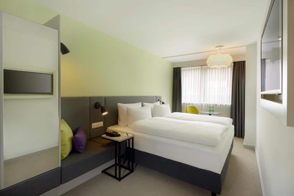 Quarto no H+ Hotel Wien, com cama de casal, zona de estar, mesas laterais, TV na parede e janela grande com cortinados transparentes.