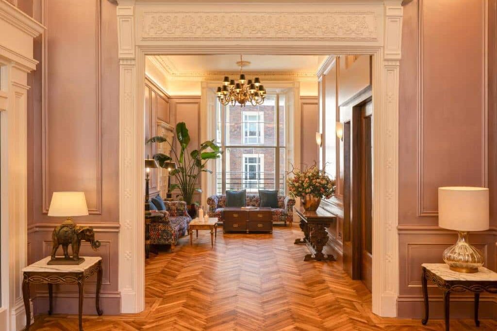 Sala de estar luxuosa no Albany House com soalho de madeira em espinha, mobiliário clássico florido, plantas grandes e lustres dourados, num ambiente opulento de estilo vitoriano.