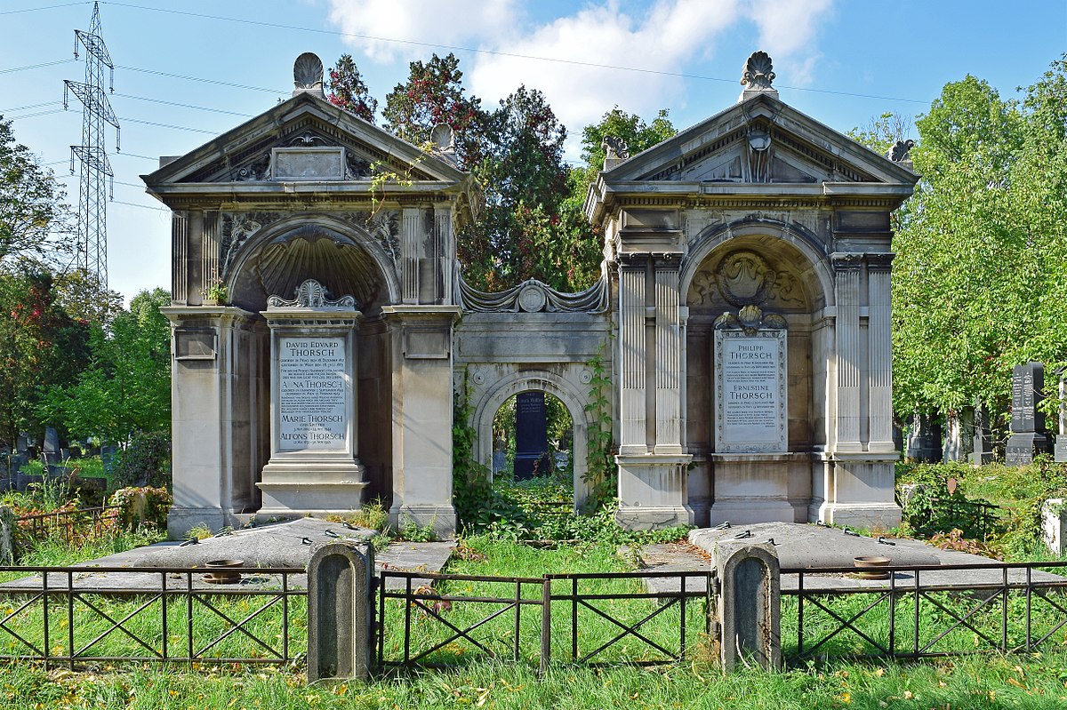 Dois mausoléus de pedra no Cemitério Central de Viena, ornamentados com arcos e inscrições, rodeados de relva e árvores.