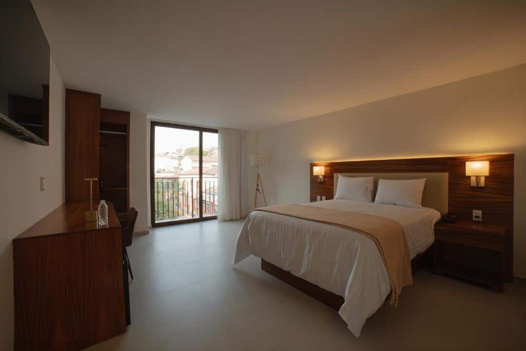 Um quarto do Hotel Casa Kavia Vallarta com uma cama de casal, móveis de madeira, secretária, TV e uma porta de vidro de correr para uma varanda.