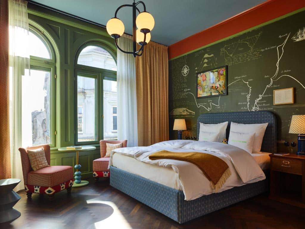 Quarto do 25hours Hotel Indre By com cama de casal, duas pequenas poltronas, mesa, candeeiro e uma parede toda desenhada. O quarto é colorido e tem duas janelas com vista para a cidade.