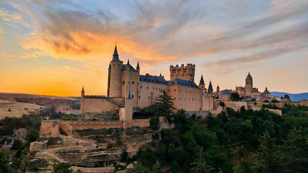 Alcázar de Segóvia, na Espanha, captado durante o pôr do sol. O castelo, com torres pontiagudas e muralhas robustas, destaca-se num penhasco. Ao fundo, o céu está laranja e rosa, criando um contraste com a estrutura histórica. Há ainda uma catedral gótica ao fundo, a acrescentar profundidade ao cenário e a realçar a história e a arquitetura da região.
