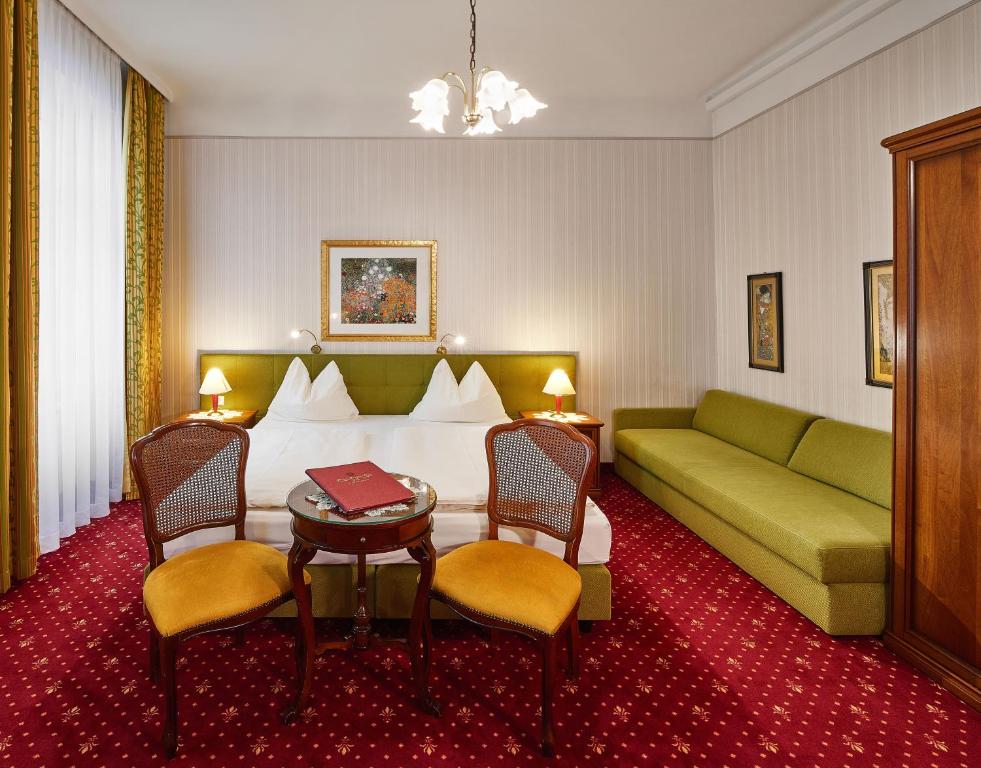 Quarto no Hotel Austria – Wien, com cama de casal, sofá verde, duas cadeiras, uma pequena mesa e obras de arte emolduradas nas paredes.