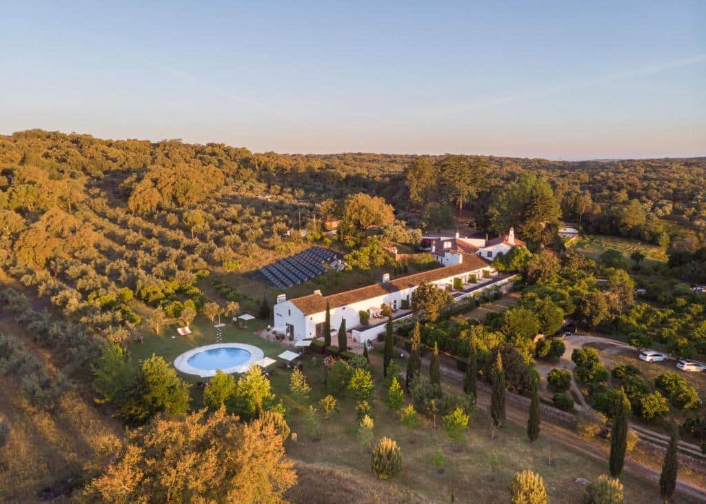 Propriedade do Imani Country House, rodeada de muito verde, árvores e colinas, com uma construção branca de telhado castanho ao centro e uma piscina redonda em frente