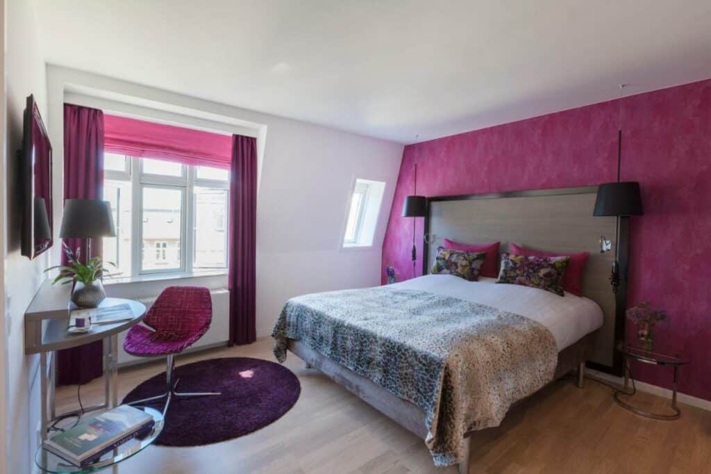 Quarto do Andersen Boutique Hotel com cama de casal, televisão e uma mesa com cadeira. Uma das paredes e os cortinados são cor-de-rosa.