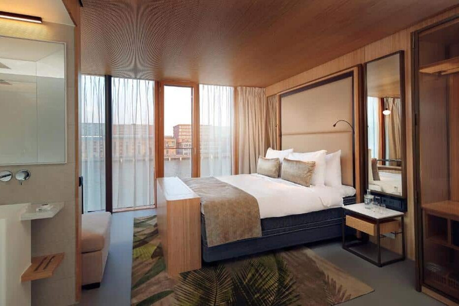 Quarto moderno do Hotel Jakarta Amsterdam, que ilustra onde ficar em Amesterdão: cama grande, pormenores em madeira, janelas do chão ao teto e vista para os edifícios em redor.