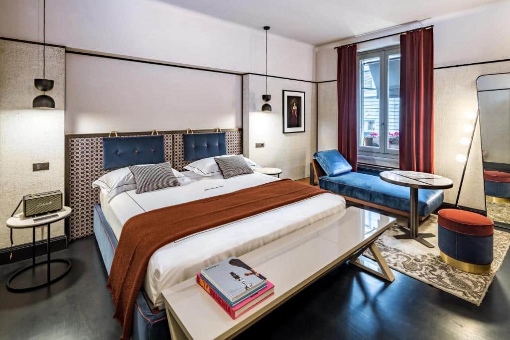 Quarto do Locanda Pandenus Brera com uma cama de casal, uma janela com cortinas, um banco aos pés da cama com alguns livros, um sofá-cama e um espelho de corpo inteiro