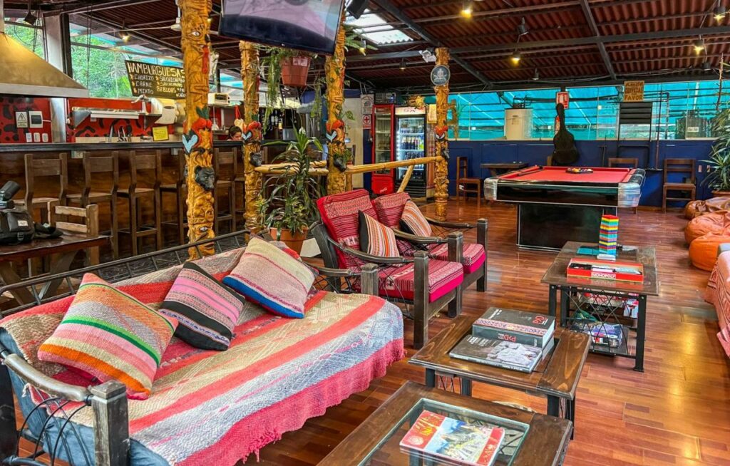 Sala comum de hostel em Machu Picchu com sofás, pufes e cadeiras, mesa de bilhar ao fundo e uma zona de bar