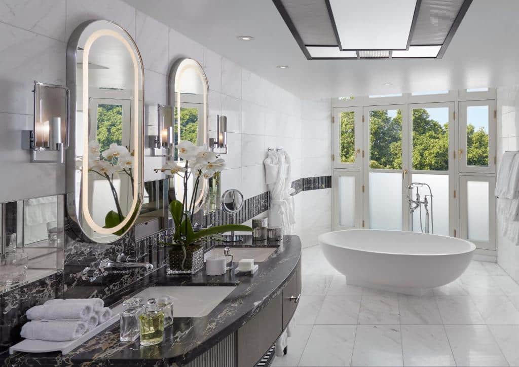 Casa de banho do Mandarin Oriental Hyde Park com banheira oval junto a uma varanda com portas de vidro, um lavatório com duas cubas e um vaso de flores.