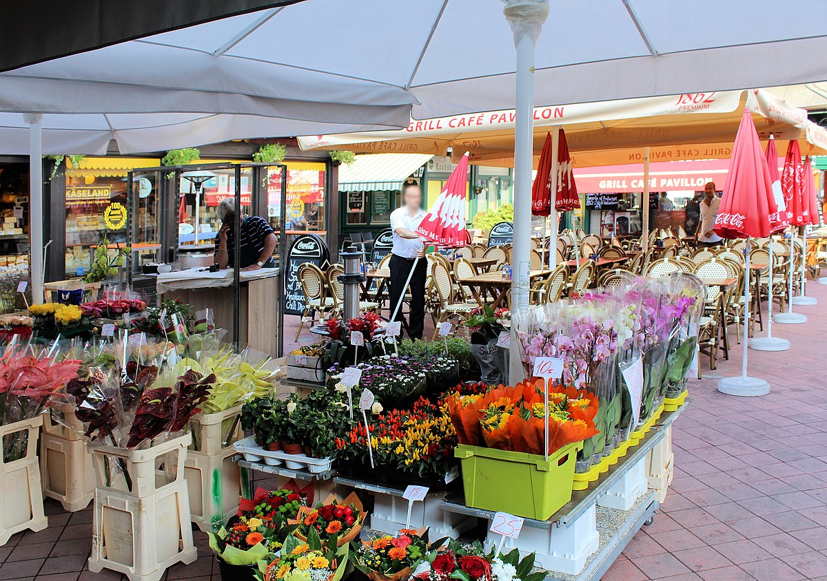 Naschmarkt, mercado ao ar livre com bancas de flores em baldes, sob grandes guarda-sóis brancos, e um café ao fundo.