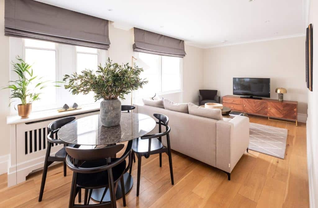 Sala de estar do Native Mayfair com duas janelas com estores, um móvel com televisão, um sofá com almofadas, uma poltrona, uma mesa redonda de quatro lugares e vasos de plantas.