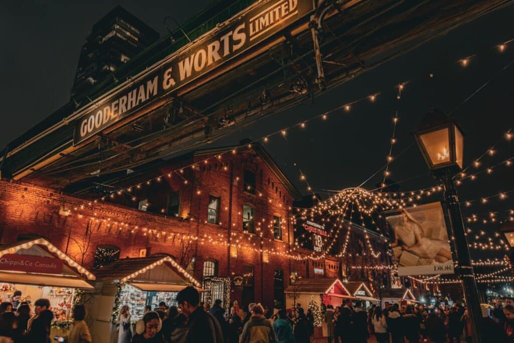 Multidão numa feira de Natal à noite, com decoração de luzes coloridas sobre construção de tijolo antigo