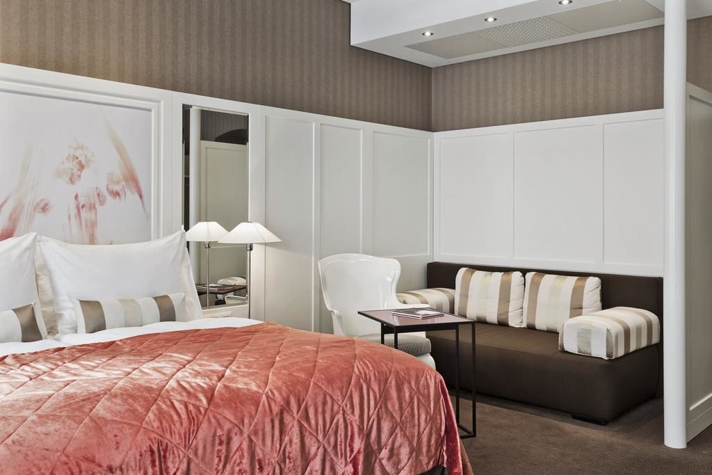 Quarto no The Harmonie Vienna, com uma cama de colcha vermelha, um pequeno sofá, uma cadeira branca, uma mesa lateral e arte moderna na parede.