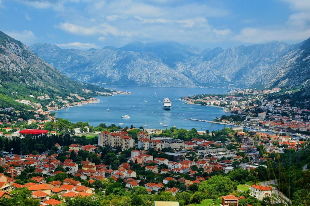 Vista panorâmica de Kotor, em Montenegro, mostrando uma baía rodeada por montanhas. A cidade é composta por numerosos edifícios com telhados de telha vermelha e vegetação verde abundante. No centro da baía, um grande navio de cruzeiro está ancorado, rodeado pelas águas e outras pequenas embarcações.