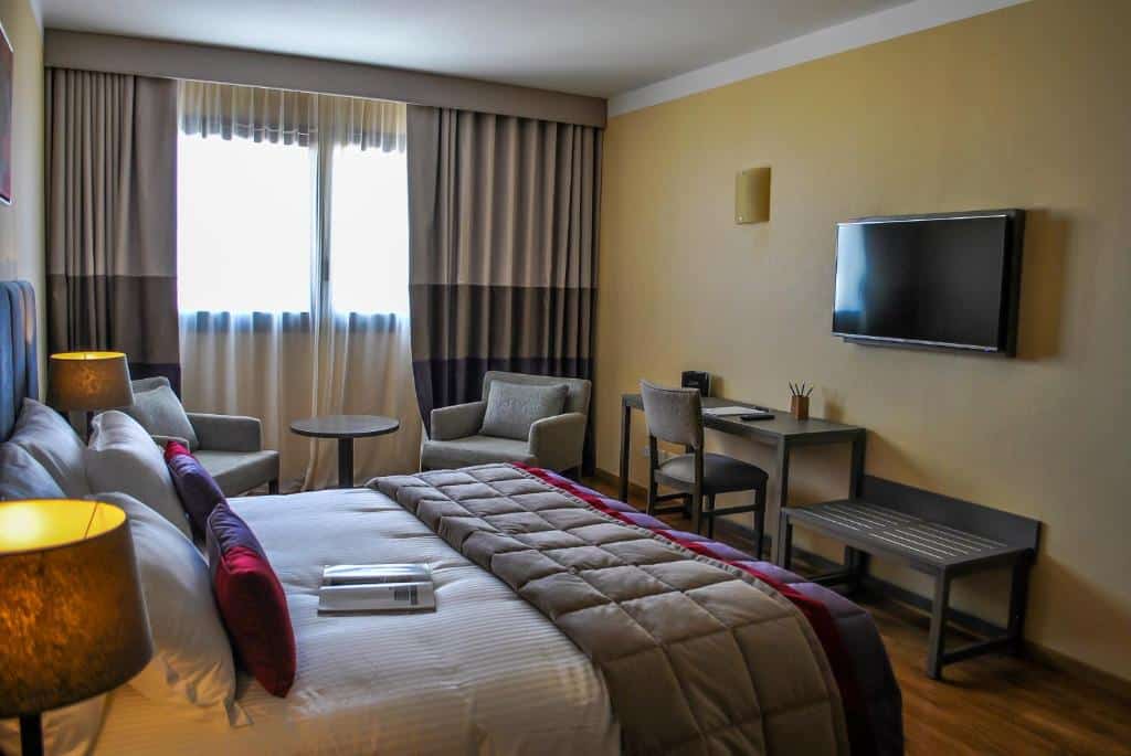 quarto do Hotel Grand Brizo Buenos Aires com cama de casal, tv e secretária com cadeira em frente, suporte para malas, mesa com duas poltronas e janela grande com cortinas