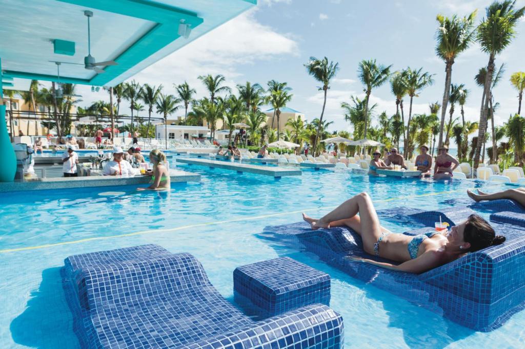 Piscina do resort com tudo incluído Riu Playacar, com espreguiçadeiras azuis e bar no meio da piscina, em dia ensolarado.