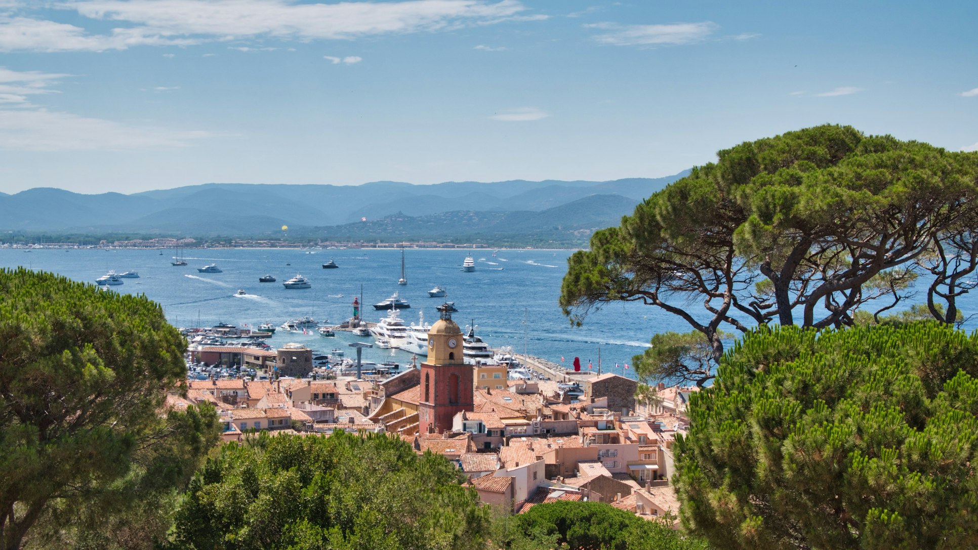 Vista de Saint-Tropez, com telhados de terracota, uma torre de igreja, barcos no porto e montanhas ao fundo.