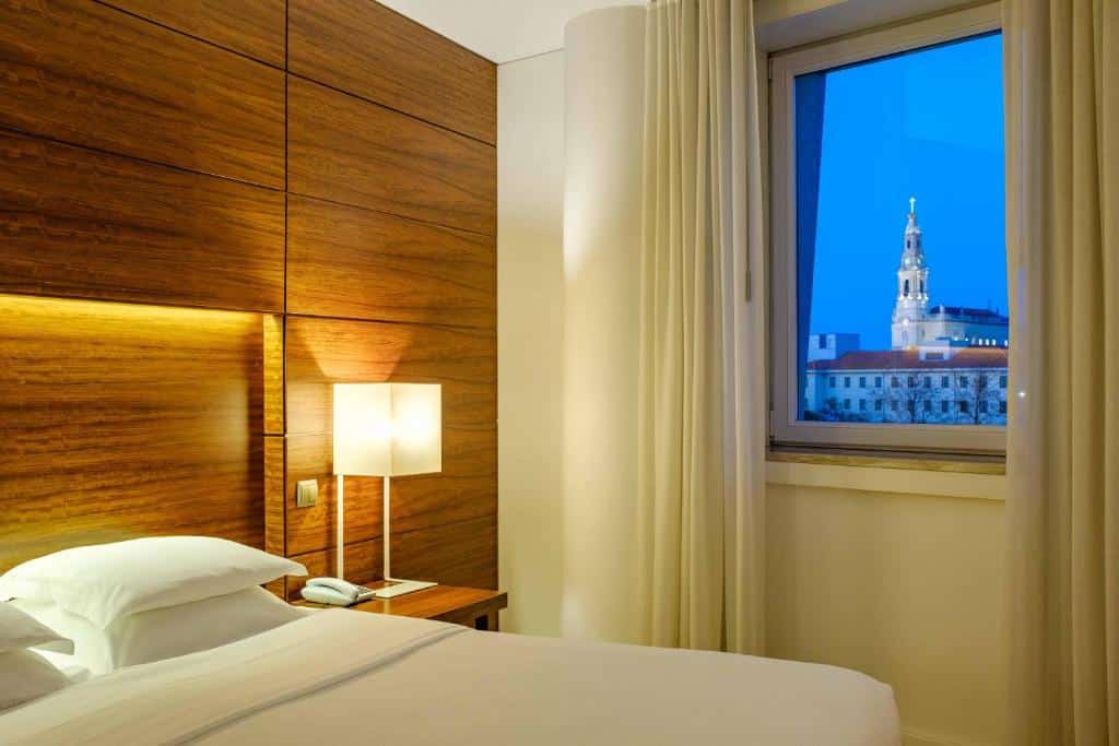 Quarto no Hotel Santa Maria com painéis de parede em madeira, candeeiro aceso, cama e uma janela com vista para a cidade e uma torre ao anoitecer.