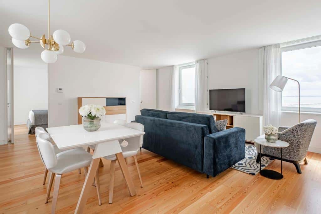 Sala de estar espaçosa do Tandem Palacio Alfama Suites com um sofá, uma poltrona, duas janelas e uma televisão; atrás de um dos sofás há uma mesa com quatro lugares e um candeeiro de teto, com pavimento que imita madeira
