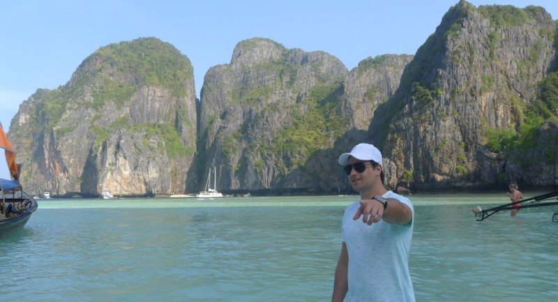 Pessoa de pé em águas rasas, com grandes penhascos rochosos e barcos ao fundo, nas ilhas Phi Phi.