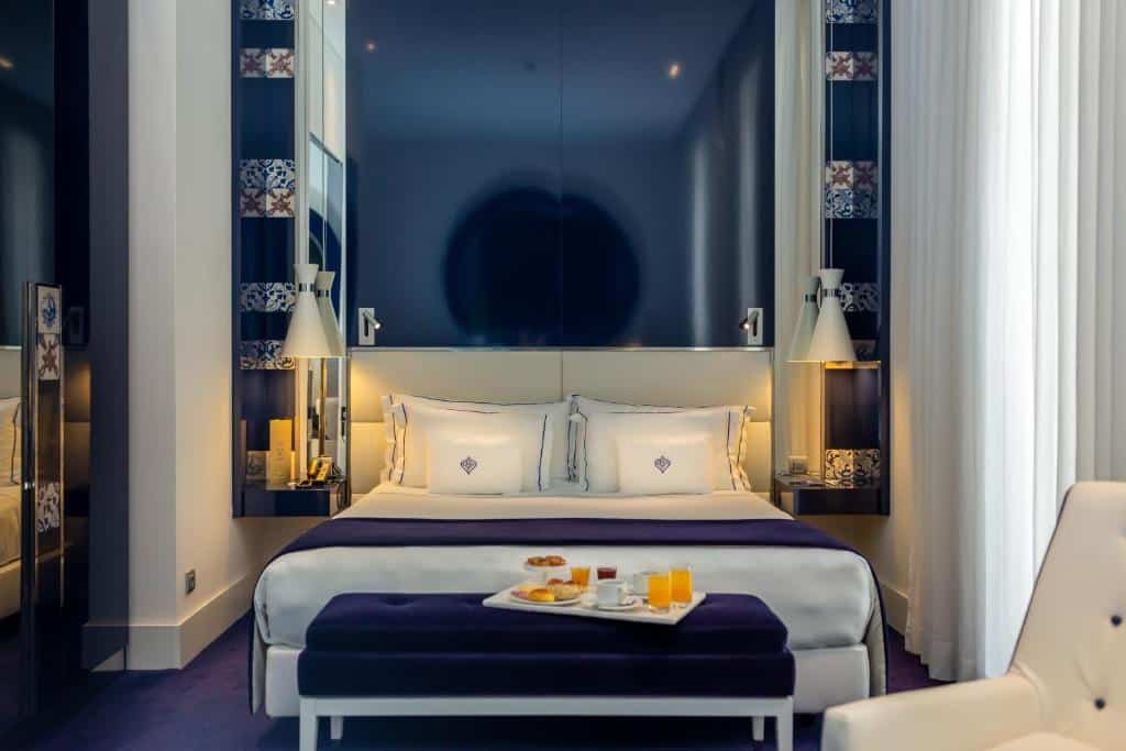Quarto do Portugal Boutique Hotel com uma cama de casal, janela com cortinas, uma poltrona branca e um banco aos pés da cama com um tabuleiro de pequeno-almoço.