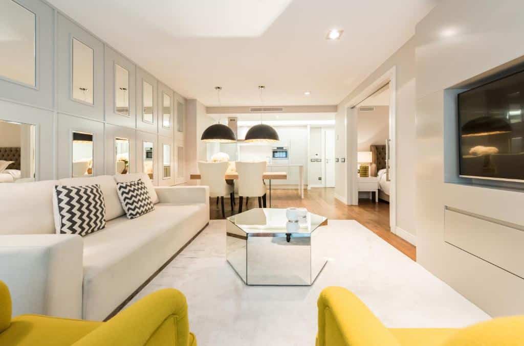 Interior de um apartamento do Gran View Apartments. A fotografia foi tirada na sala, onde um sofá branco e duas poltronas amarelas estão viradas para uma mesa espelhada e uma TV na parede. Ao fundo está a cozinha, com mesa de jantar e cadeiras. A parede ao lado está cheia de espelhos e, junto à cozinha, uma porta dá acesso ao quarto.