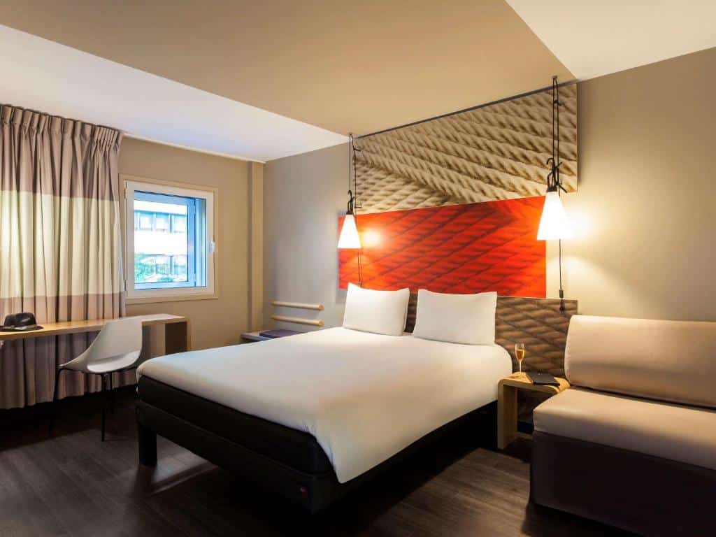 Quarto do Ibis Lisboa Liberdade com uma cama de casal, uma janela com cortinas, uma secretária com cadeira e um pequeno sofá com um candeeiro ao lado