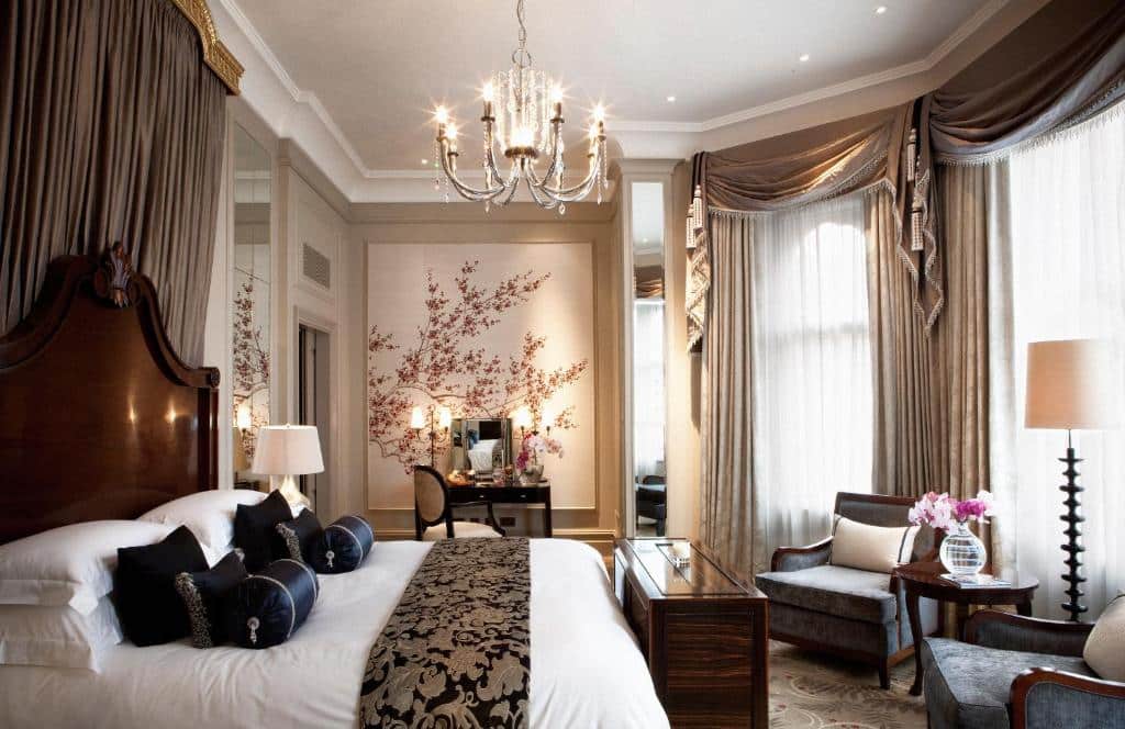 Quarto luxuoso do The Langham London com janelas com cortinas, um lustre, cama de casal com muitas almofadas, um toucador, duas poltronas e um candeeiro de pé.