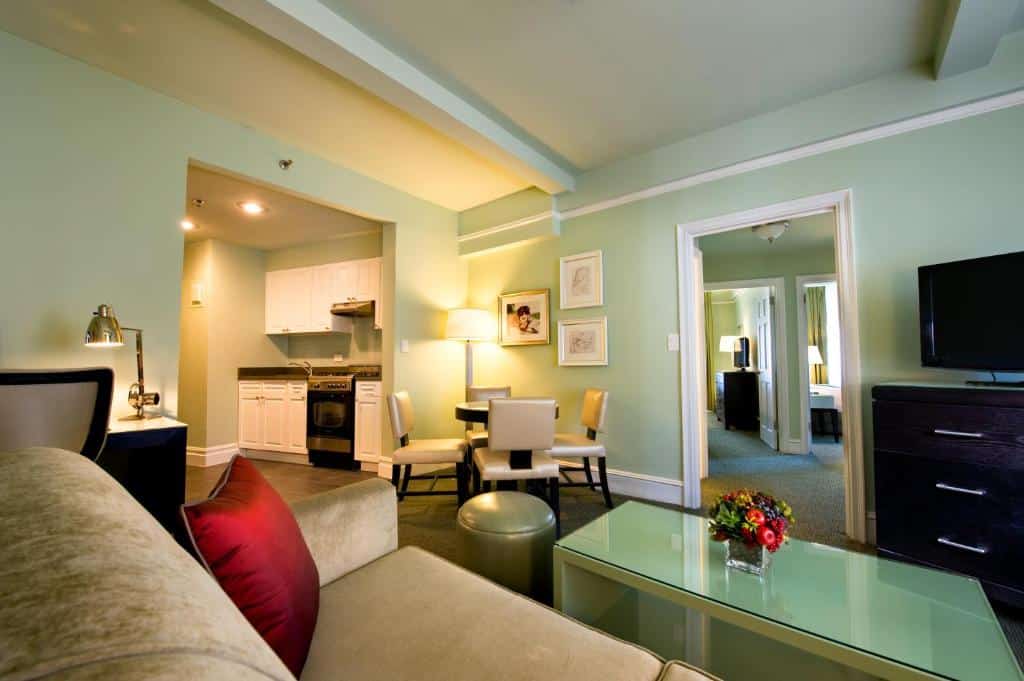 Sala de estar de um hotel com sofá, mesa de jantar e cozinha americana equipada. Decoração acolhedora, em tons suaves de verde, com mobiliário confortável.