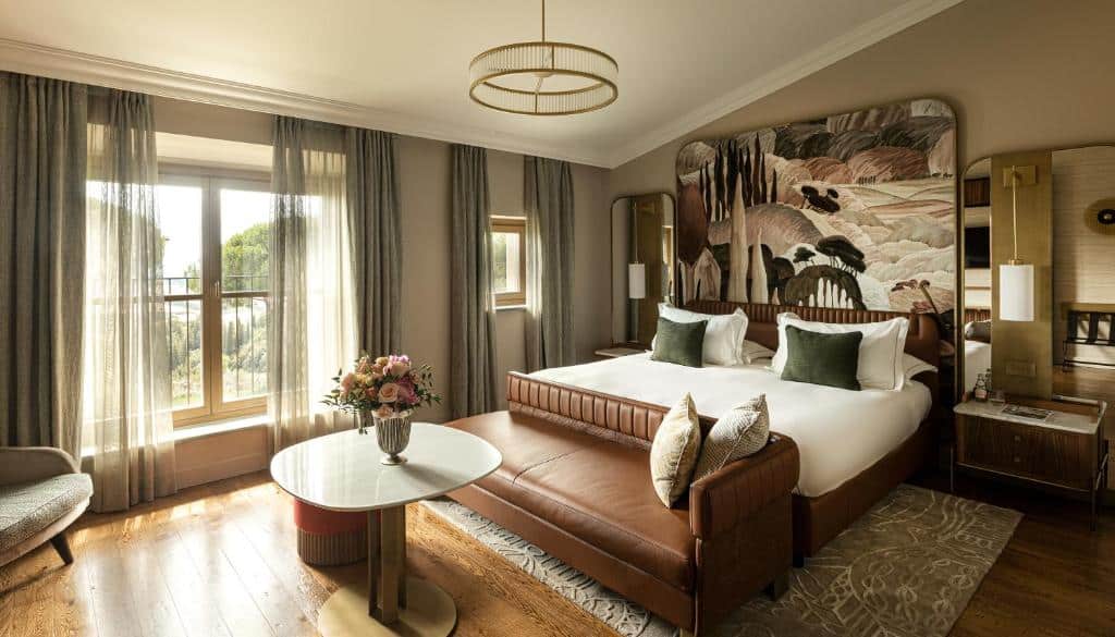 Quarto sofisticado do Toscana Resort Castelfalfi, em tons de castanho e verde-militar, com cama de casal, sofá de couro à frente, mesa com uma flor e janelas grandes com vista sobre a natureza