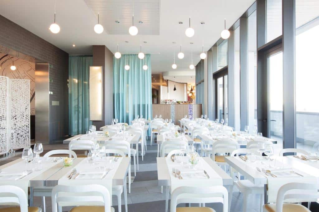 salão do Ibis Styles Roma Eur com mesas e cadeiras brancas, candeeiros em forma de bola por cima, pormenores em verde-água e um grande balcão ao fundo