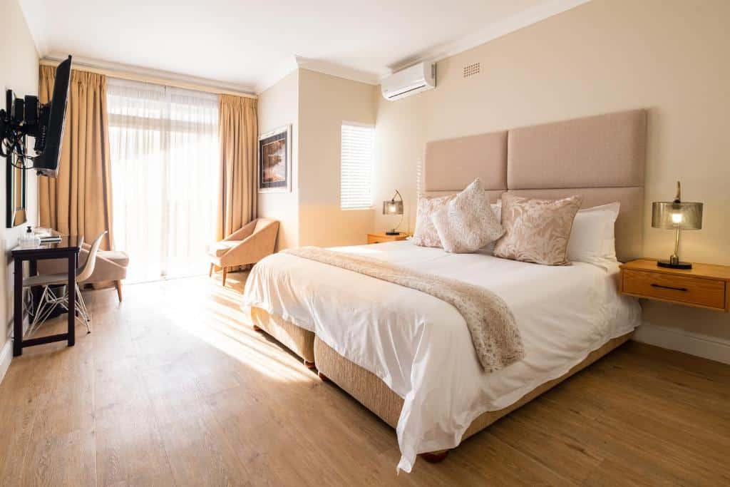 Quarto bem iluminado do 3 On Camps Bay com cama grande, mesas laterais, secretária, cadeira, TV e janela do chão ao teto.