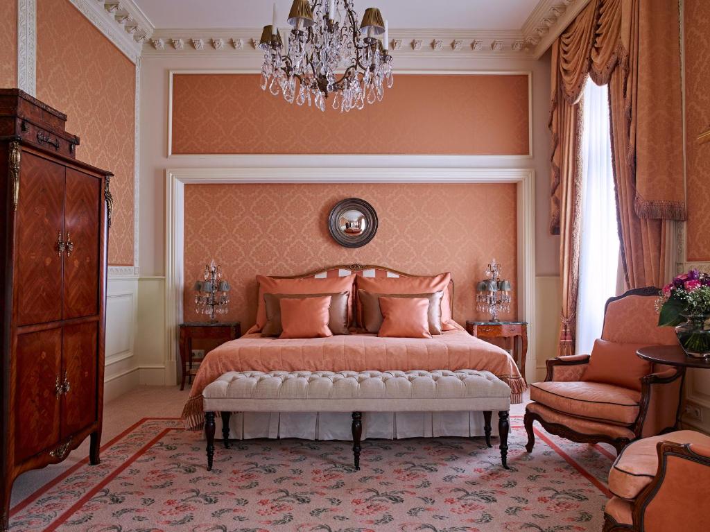 Quarto elegante no Grand Hotel Wien, com paredes cor de pêssego, um lustre, uma cama de casal, poltrona e mobiliário de madeira ornamentado em estilo clássico.
