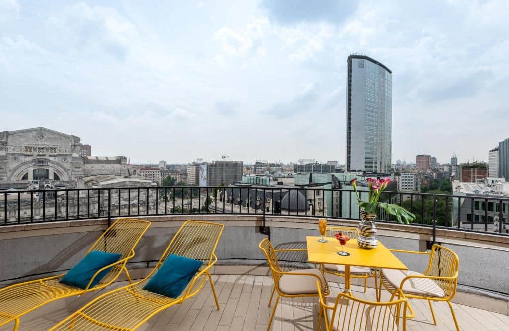 Terraço na cobertura do NYX Hotel Milan com vista para a estação central e outros edifícios, com espreguiçadeiras, mesa e cadeiras de ferro amarelo